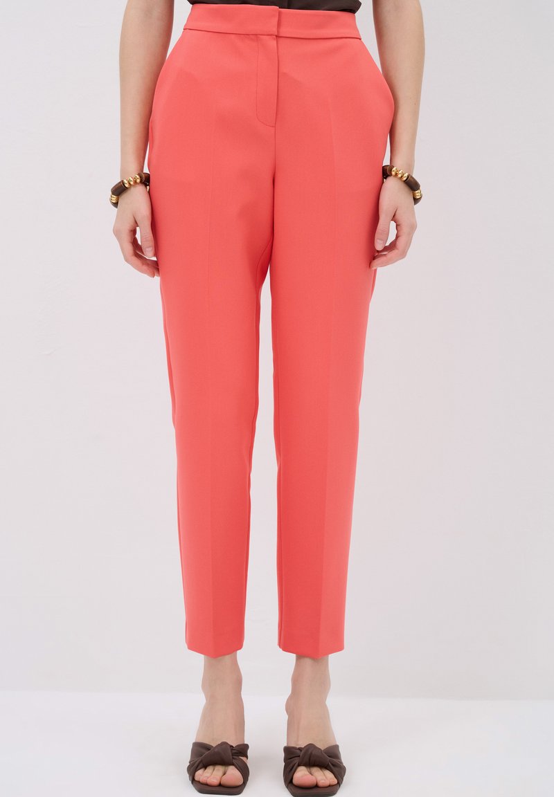 Coral tailored trousers with a slim fit, featuring side pockets and sharp creases down the front. Paired with brown shoes.