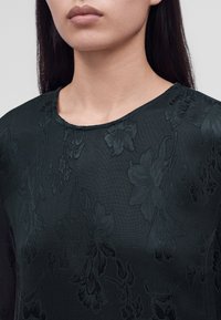 Femme aux cheveux raides et foncés porte un haut sombre en tissu à texture florale avec un col rond, vu du menton aux épaules sur un fond clair.