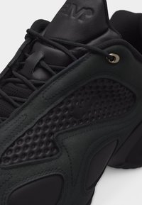 Közeli kép egy fekete tornacipőről, melynek oldalpanelei textúráltak, fekete fűzője van, a bőrből készült nyelvén dombornyomott "AIR" felirat található, és egyedi talpkialakítása van.