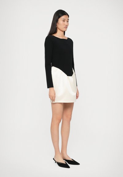 Pikkade mustade juustega naine, kes kannab musta ja valget mini kleiti ning musti terava ninaga slingback jalatseid, seisab lihtsa valge tausta ees.