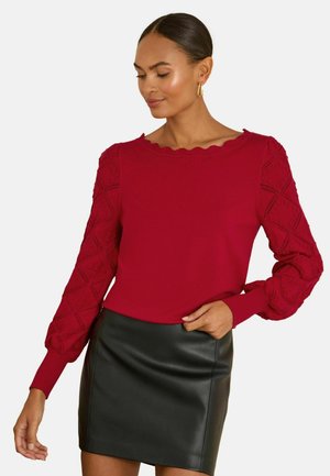 Red sweater featuring textured, puffed sleeves with diamond patterns and a scalloped neckline, paired with a black leather skirt.