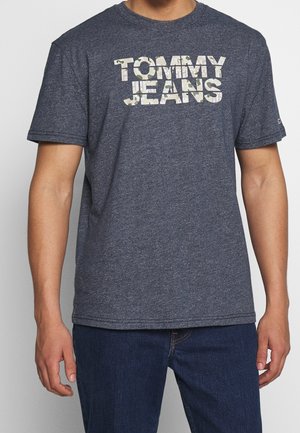 Man draagt een blauw T-shirt met korte mouwen met de tekst "TOMMY JEANS" in camouflagepatroon over de borst, gecombineerd met donkerblauwe jeans.