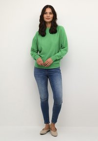 Groene gebreide trui met lange mouwen, ronde hals, gecombineerd met strakke blauwe jeans en beige platte schoenen. Soepele textuur, casual ontwerp.