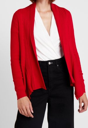 Femme portant un haut blanc en col V, un cardigan rouge ouvert côtelé et un pantalon noir taille haute, debout devant un fond uni.
