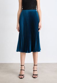 Gonna midi plissettata in teal realizzata in tessuto morbido, con vita aderente e silhouette fluida. Sandali neri con tacchi a stiletto completano il look.