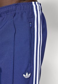 Zilas sporta bikses ar baltām trim svītrām, kurās ir rāvējslēdzēja kabata un mazs balts logotips uz kreisās kājas.