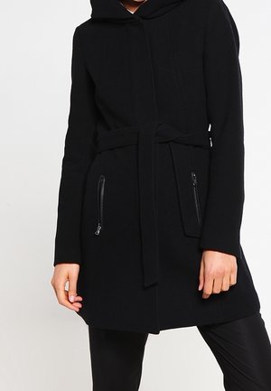 Person iført en sort hættejakke med bælte om livet og to lynlåslommer foran, stående mod en ensartet lys baggrund.