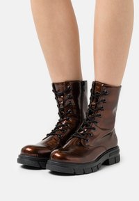 Bottines en cuir verni marron à lacets avec une semelle en caoutchouc noire épaisse, dotées de lacets noirs et d'une texture lisse et brillante.