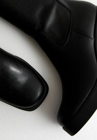 Botas de tobillo de cuero negro con una textura suave, punta redondeada y sutilezas en la costura. El diseño presenta un tacón bajo y robusto.