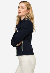 Chaqueta azul marino con un diseño ajustado, que presenta cremalleras blancas y acentos verdes. Textura suave con cuello vuelto y corte entallado.