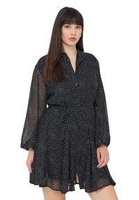 Abito nero con pois bianchi, tessuto leggero e trasparente, maniche lunghe, chiusura frontale con bottoni, cintura in vita e orlo a strati con rouches.
