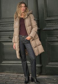 Geacă matlasată bej cu glugă, design matlasat și patch cu logo, asortată cu blugi din denim închis și cizme negre cu toc. Textură și stil modern.