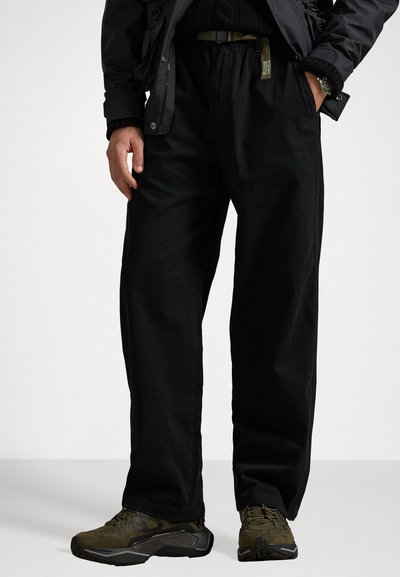 Pantaloni neri, morbidi e larghi con vita elasticizzata e due tasche laterali, realizzati in un tessuto resistente. Indossati con scarpe scure caratterizzate da dettagli testurizzati.