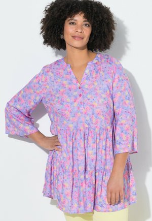 Femme aux cheveux bouclés portant une tunique violette à motifs floraux pastel avec des manches trois-quarts, debout devant un fond blanc, la main sur la hanche.