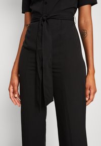 Combinaison noire à manches courtes, avec une taille nouée et un tissu lisse. Lignes épurées et coupe ajustée.