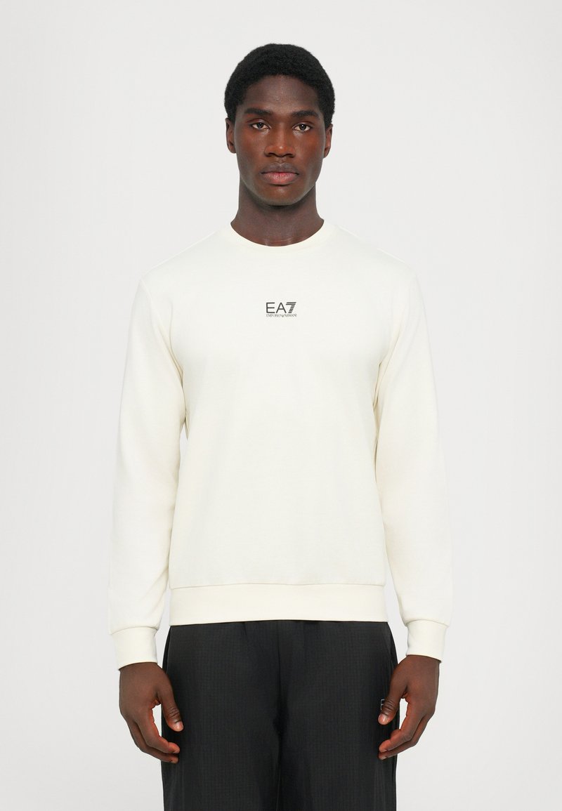 Sudadera color crema con cuello redondo y mangas largas. Presenta un pequeño logo negro de EA7 en el pecho. Tejido suave y texturizado.