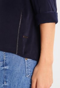 Närbild av en person som bär en marinblå skjorta med uppkavlade ärmar och synlig sömdetalj, tillsammans med ljusblå denimjeans ficka.