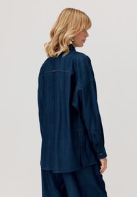 Kobieta z blond falującymi włosami w granatowej, oversize'owej dżinsowej kurtce i pasujących spodniach, stojąca bokiem na jednolitym tle.