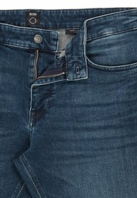 Tmavě modré džínové kalhoty s průhmatovým zapínáním na zip, viditelným šitím a výrazným designem kapes. Materiál vykazuje mírné opotřebení a variace textury.