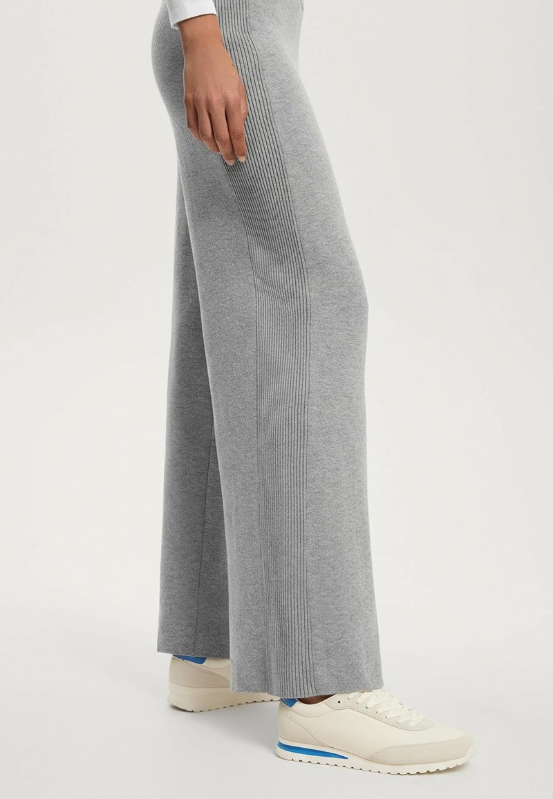 Szürke, bő szárú nadrág bordázott oldalsó mintázattal, puha anyagból készült. Krémszínű tornacipővel párosítva, amelyen kék díszcsík található.