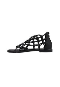 Sandalias negras con un diseño de tira trenzada, suela plana, textura suave y cierre con cremallera en la parte posterior. Estructura minimalista y abierta.