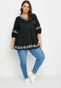 Top in cotone nero con scollatura a V e maniche a palloncino, accentuato da ricami floreali bianchi lungo l'orlo e i polsini, abbinato a jeans blu.