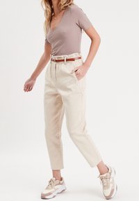 Pantalon beige taille haute avec ceinture marron, présentant une coupe décontractée et des poches latérales. Associé à des baskets beige et blanches. Haut à manches courtes.