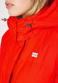 Rode geïsoleerde jas met hoge kraag, verstelbare trekkoorden en een Levi's logo patch op de borst. Gladde textuur, afgeronde vorm.