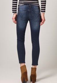 Osoba v tmavě modrých skinny džínách a hnědých semišových kotníkových botách na vysokém podpatku, stojící na jednobarevném světlém pozadí.