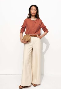 Chemise à manches longues de couleur rouille avec des boutons, associée à un pantalon crème à jambes larges. Tient un sac en beige tissé texturé. Ceinture marron et sandales plates.