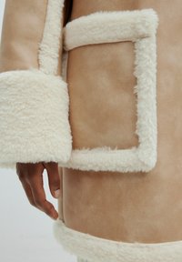 Bruine leren buitenkant met een fluffy crèmekleurige voering. Bevat een grote patchzak en zachte, gestructureerde manchetten, die hoge-contrasterende materialen tonen.