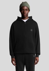 Felpa nera con zip e tasca frontale, completa di patch logo, indossata sopra una maglietta bianca. Il modello indossa anche un berretto lavorato a maglia verde.