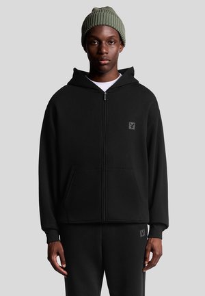 Schwarze Zip-Hoodie mit einer Fronttasche und einem Logopatch, über einem weißen Shirt getragen. Das Modell trägt außerdem eine grüne Strickmütze.
