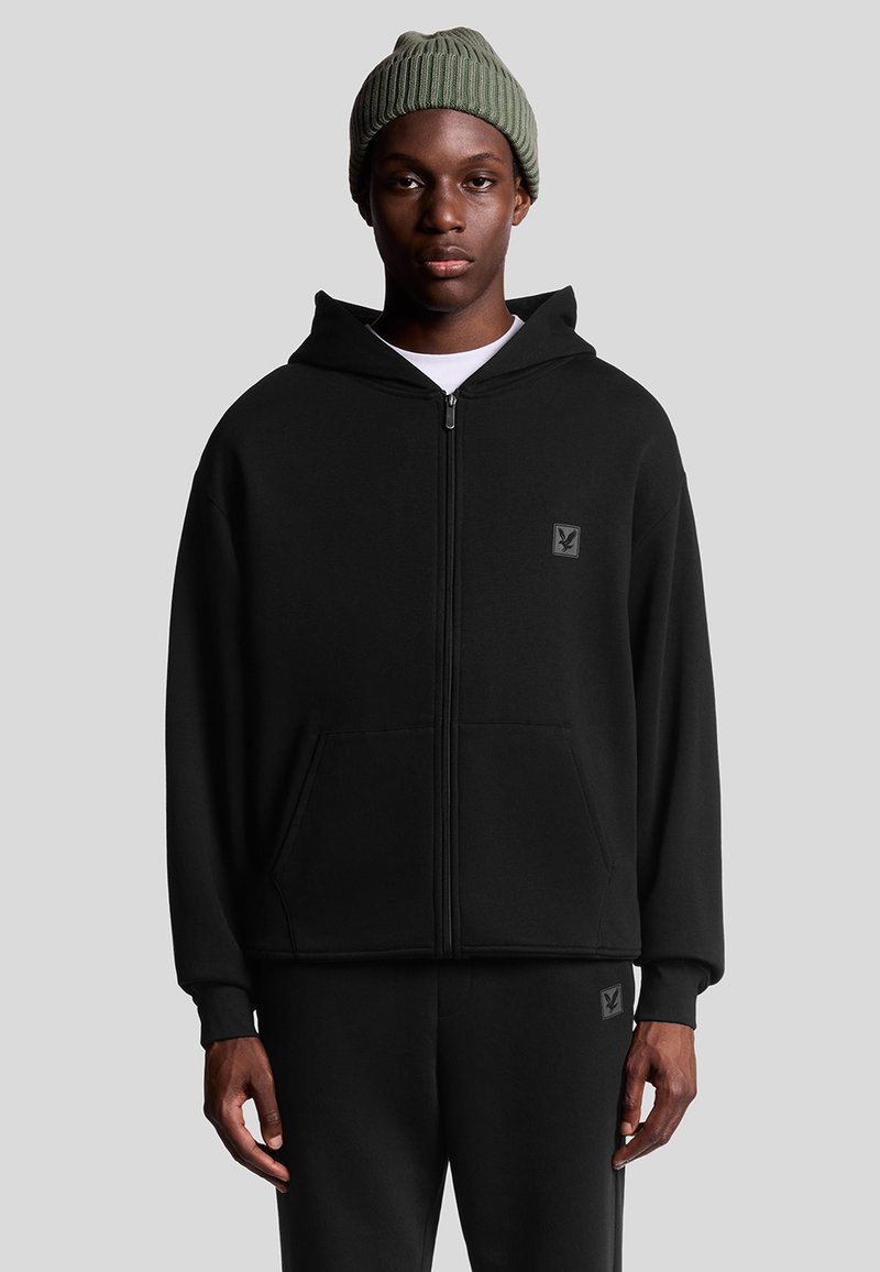 Felpa nera con zip e tasca frontale, completa di patch logo, indossata sopra una maglietta bianca. Il modello indossa anche un berretto lavorato a maglia verde.