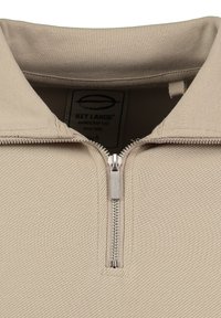 Sudadera de color beige con cremallera, hecha de una tela texturizada, que cuenta con un cuello alto y una cremallera de metal. La etiqueta interior dice "Key Largo, hecho a mano."