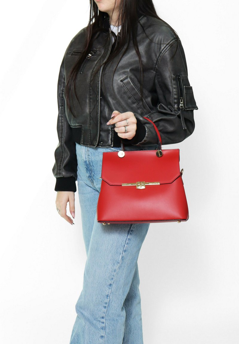 Borsa a mano rossa con design strutturato, materiale liscio, dettagli in oro e manico superiore, abbinata a una giacca di pelle nera e jeans blu.