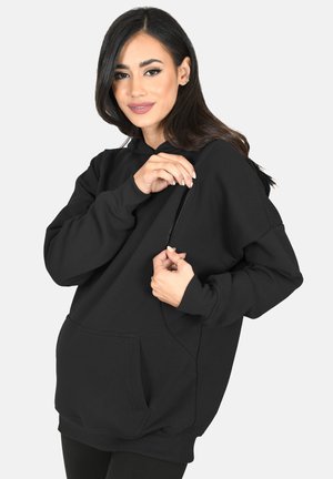 Woman with long dark hair wearing black hoodie with a zippered side pocket, smiling and holding the zipper open with both hands.