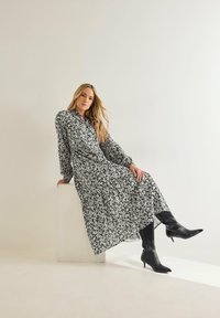 Schwarzes und weißes Blumenkleid mit langen Ärmeln und gestuftem Rock, kombiniert mit enganliegenden schwarzen Stiefeln bis zum Knie, vor einem neutralen Hintergrund.