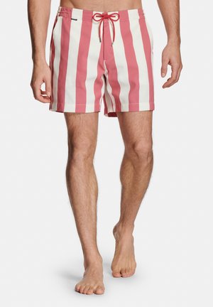 Mann trägt knielange Badeshorts mit vertikalen rosa-weißen Streifen, rotem Kordelzug in der Taille, steht barfuß vor weißem Hintergrund.