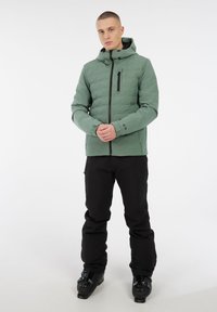 Mann in einer grünen wattierten Jacke, schwarzen Schneehosen und schwarzen Skischuhen, der vor einem weißen Hintergrund steht.