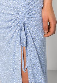 Vestido azul claro com padrões florais brancos, apresentando franzidos laterais e um detalhe de laço. O tecido é suave, com um leve brilho e uma fenda na coxa.