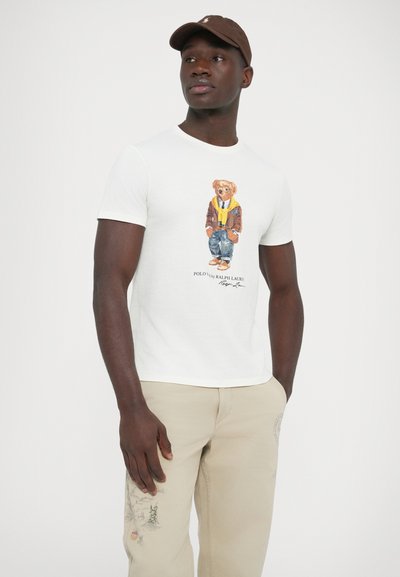 T-shirt branca de algodão com estampa de um urso vestido com uma jaqueta castanha e jeans azuis. Combinada com calças bege com detalhes bordados.