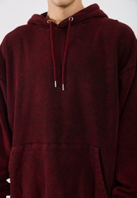 Burgundi oversized kapucnis pulóver hosszú ujjakkal, húzózsinórral és elülső zsebbel. Puha anyag finom textúrával és kontrasztos varrással.