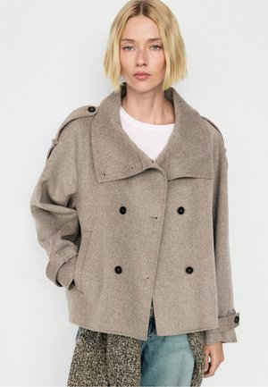Donna bionda con un cappotto di lana beige doppiopetto con bottoni neri, una maglietta bianca, jeans blu e una sciarpa fantasia.