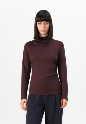 Femme aux cheveux longs et raides de couleur noire, portant un haut maroon à col montant à manches longues et un pantalon bleu marine, se tenant devant un fond blanc.