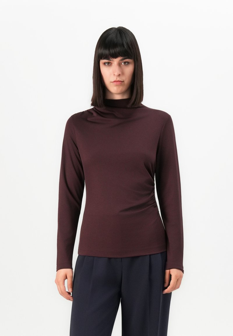 Vrouw met stijl donker haar, gekleed in een lange mouwen bordeauxrode coltrui en donkerblauwe broek, staande tegen een witte achtergrond.