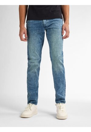 Homme debout portant un jean denim bleu, des baskets blanches et une chemise noire, visible de la taille aux pieds sur un fond blanc.
