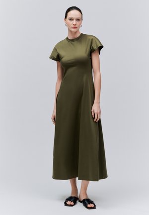 Žena stojící v olivově zelených midi šatech s krátkým rukávem a černých nazouvacích sandálech na jednoduchém šedém pozadí.