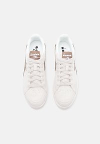 White suede sneakers with brown accents, featuring a round toe, lace-up front, and padded collar. Brand logo on tongue and insole.