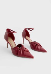 Chaussures à talons hauts à bout pointu en simili cuir bordeaux, dotées d'un design à l'avant torsadé et d'une lanière réglable à la cheville. Texture lisse.